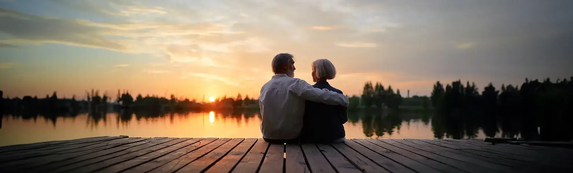 Ein Ehepaar sitzt eng umarmt auf einem See auf einen Steg und schaut sich den Sonnenuntergang an.
